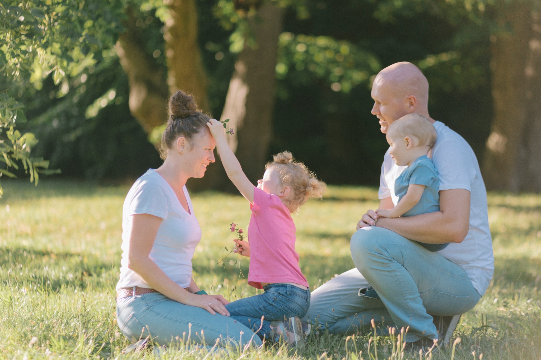 Rodzina siedząca na trawie w parku: mama z upiętymi włosami, tata trzymający synka i córka wręczająca mamie kwiatek.