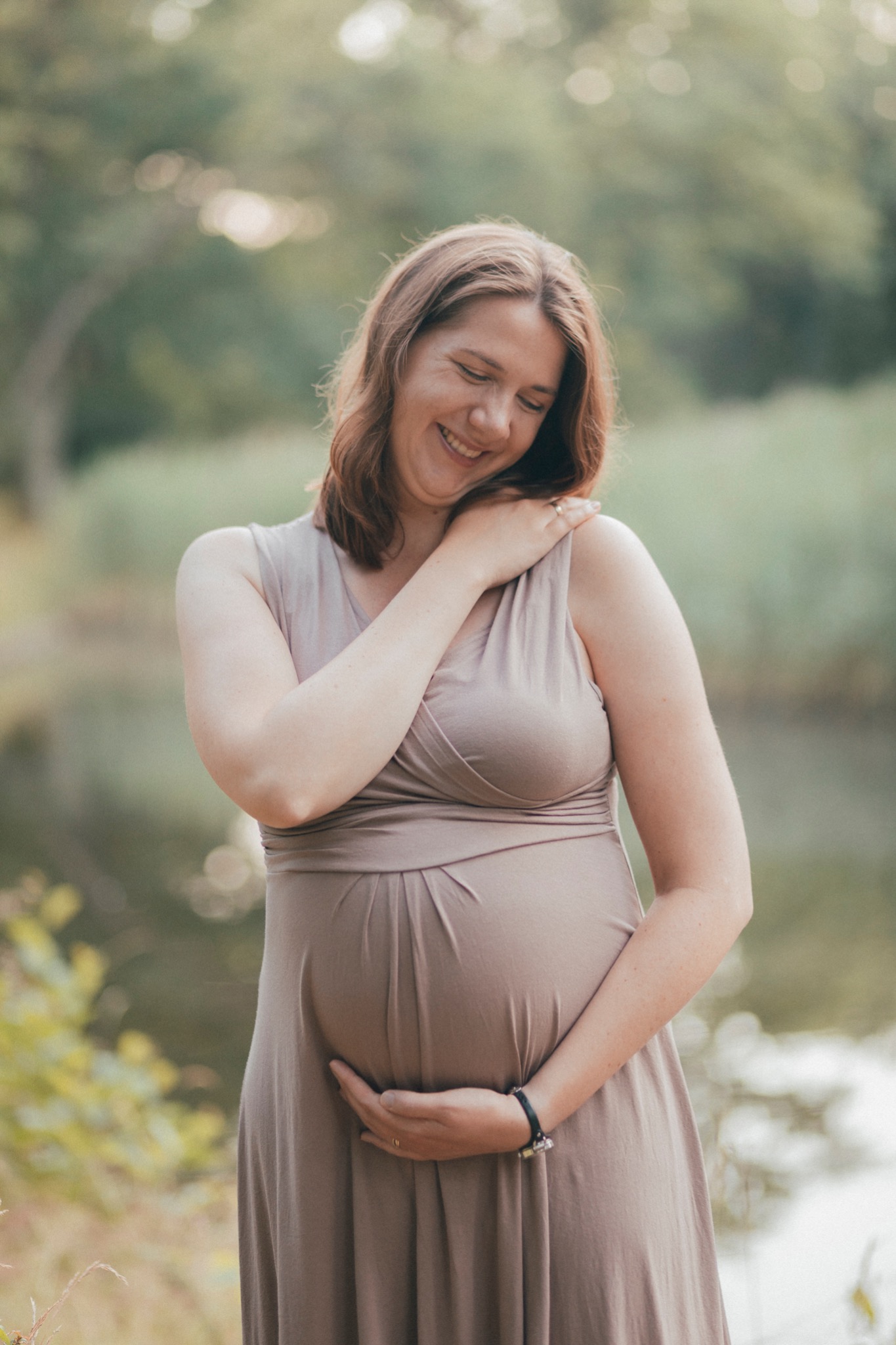 Uśmiechnięta kobieta w zaawansowanej ciąży, w beżowej sukience, pozuje na tle zieleni i wody, trzymając dłonie na brzuchu i opierając rękę na ramieniu.