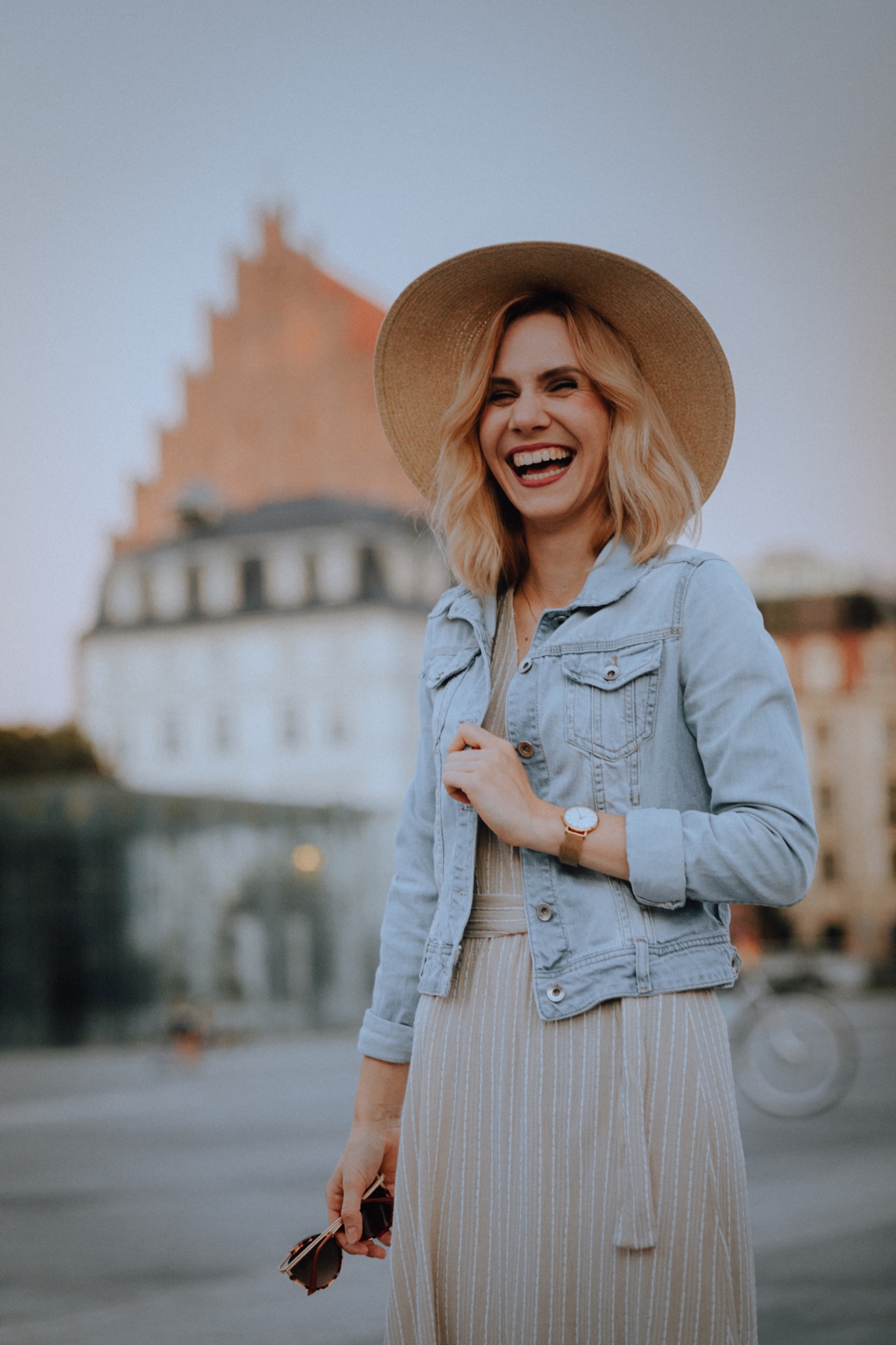 Uśmiechnięta kobieta w kapeluszu i dżinsowej kurtce na tle miejskiego budynku, trzymająca okulary przeciwsłoneczne