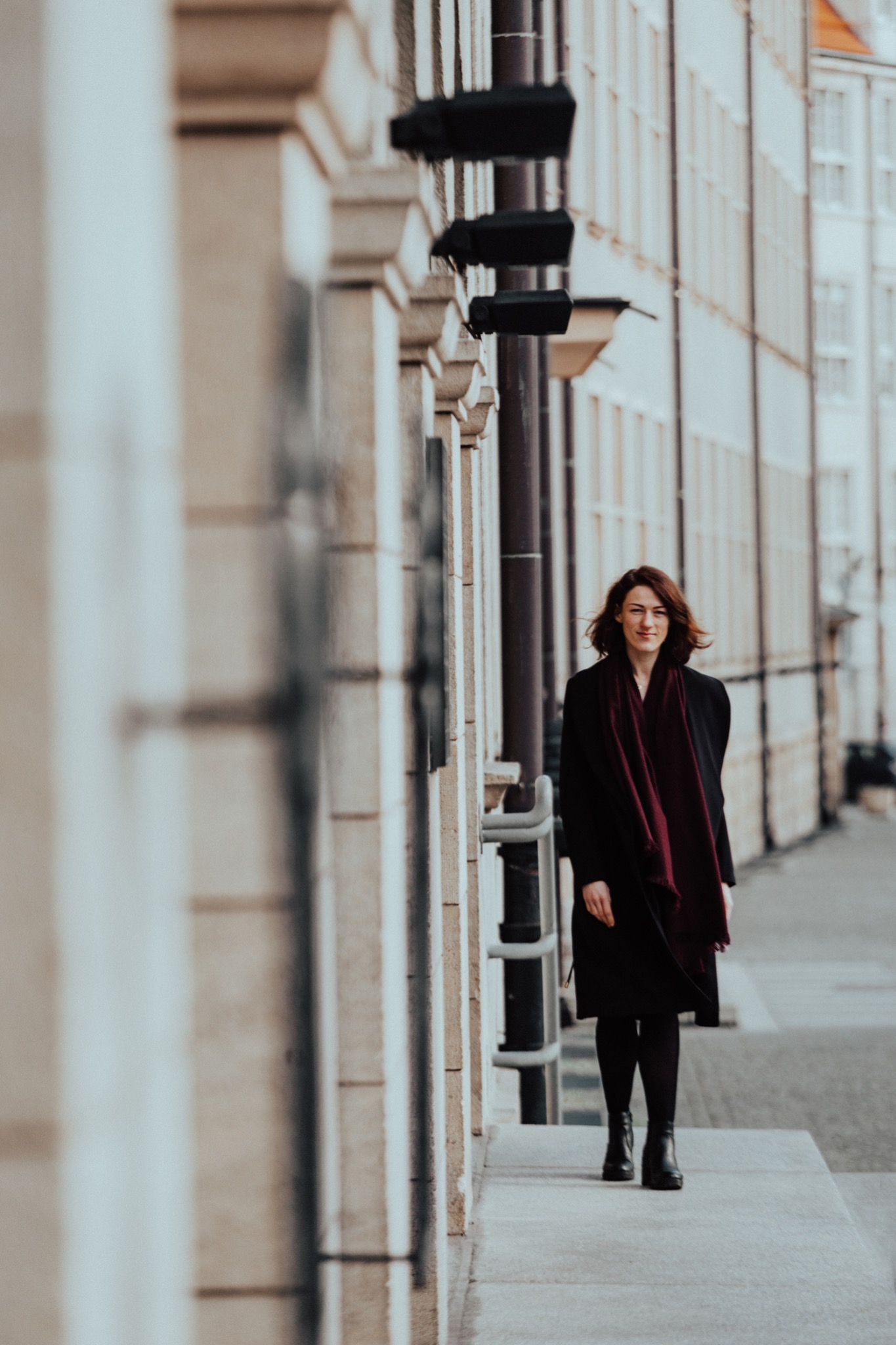 Kobieta w ciemnym płaszczu i bordowym szalu spaceruje wzdłuż budynku z kolumnami, patrząc w obiektyw.
