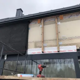 Prace na elewacji budynku z widocznym ociepleniem, folią paroizolacyjną, drewnianymi elementami konstrukcyjnymi oraz rusztowaniem. W tle widoczny krajobraz przez okno, a na pierwszym planie...