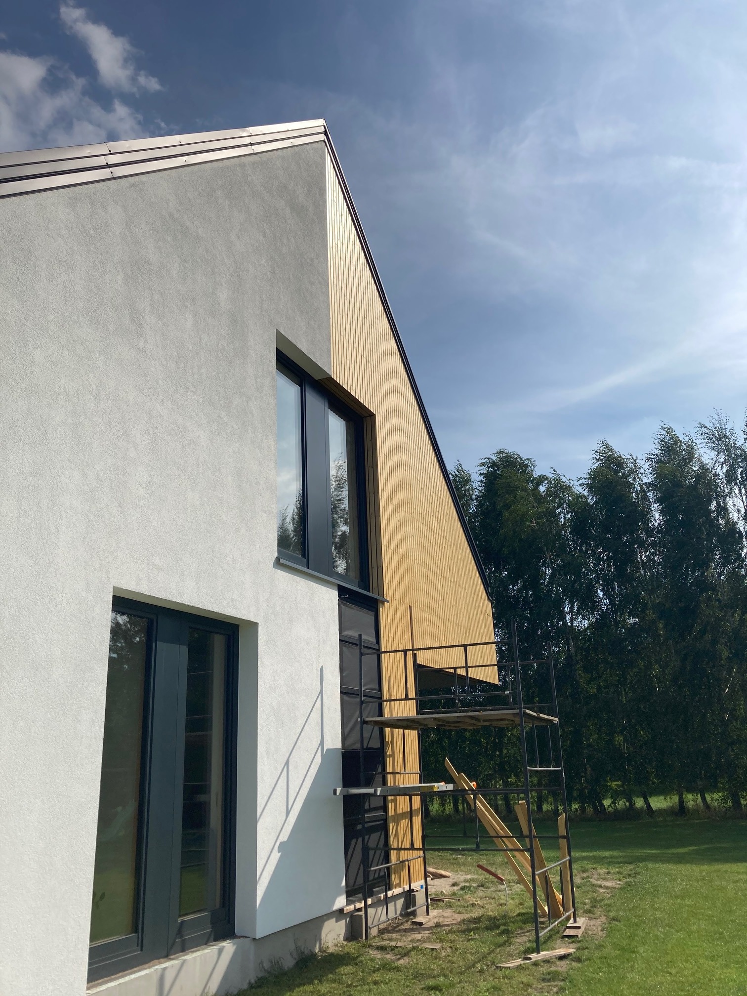 Nowoczesny dom szkieletowy z drewnianą elewacją i białym tynkiem, widoczne rusztowanie przy ścianie bocznej, błękitne niebo w tle.