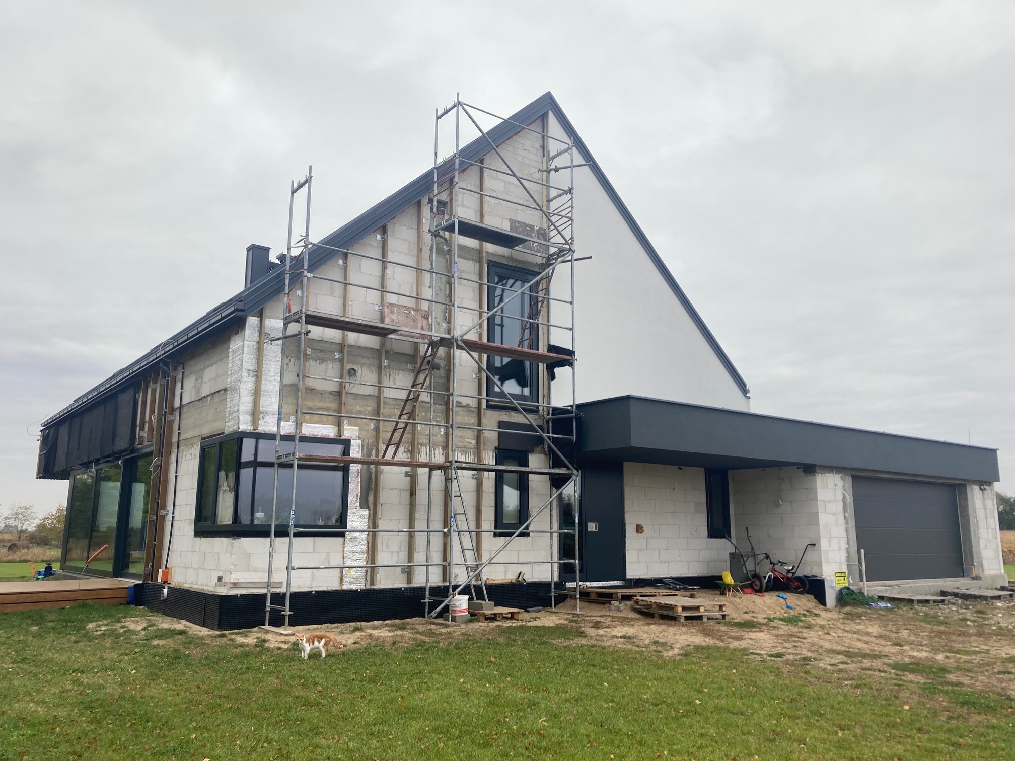 Nowoczesny dom szkieletowy w trakcie budowy, widoczne rusztowania, ocieplenie ścian i fragment wykończonej elewacji, na pierwszym planie kot.