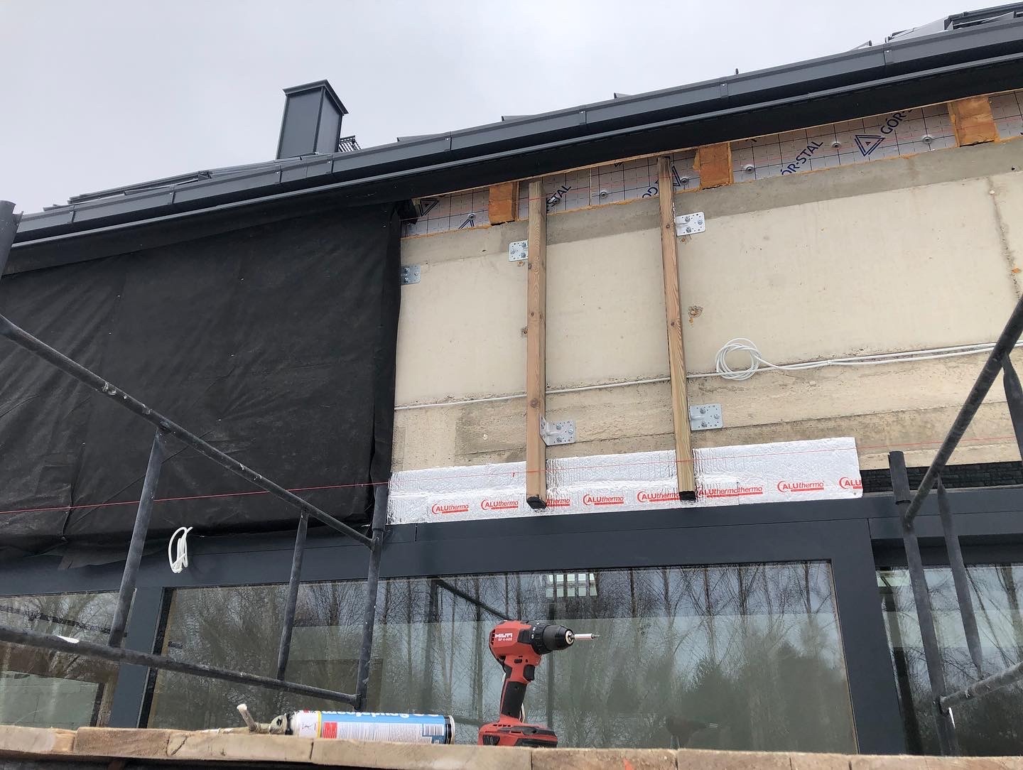 Prace na elewacji budynku z widocznym ociepleniem, folią paroizolacyjną, drewnianymi elementami konstrukcyjnymi oraz rusztowaniem. W tle widoczny krajobraz przez okno, a na pierwszym planie...