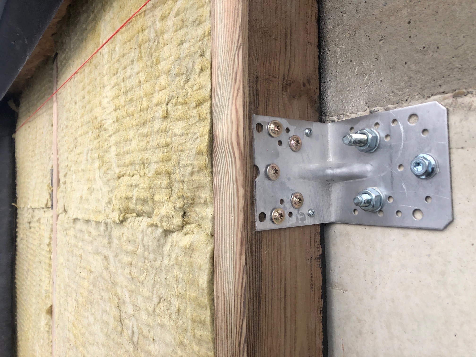 Fragment ściany w trakcie ocieplania: widoczna wełna mineralna, drewniana rama i metalowy łącznik kątowy przytwierdzony do betonowej ściany za pomocą śrub i nakrętek.