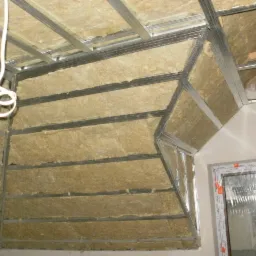 Montaż izolacji z wełny mineralnej w przestrzeni poddasza, widoczne metalowe profile konstrukcyjne i tymczasowe oświetlenie żarówką.