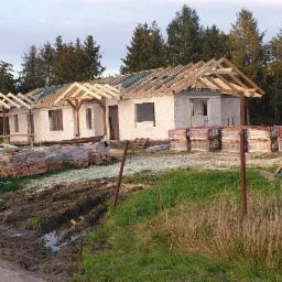 Budowa domu w stanie surowym otwartym: ściany z bloczków, drewniana konstrukcja dachu bez pokrycia, palety z dachówką obok budynku.