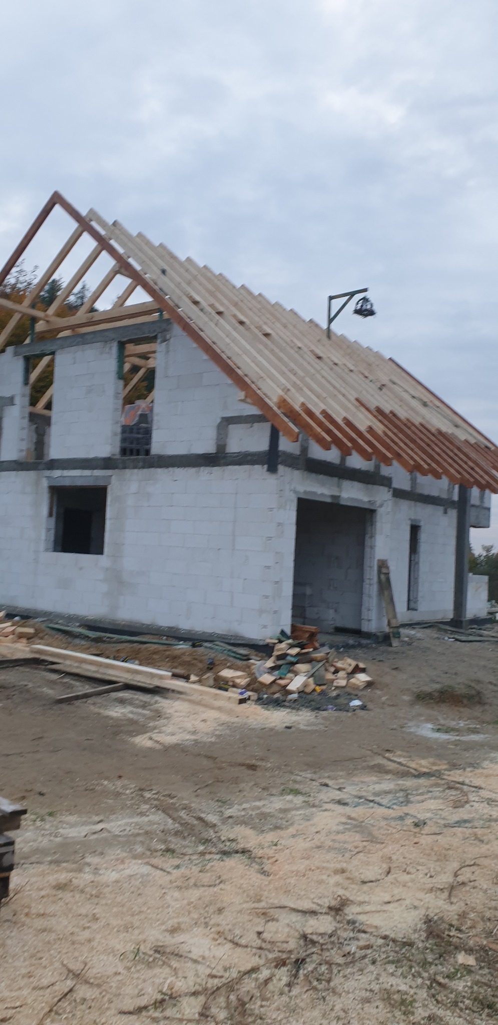 Surowy stan domu z białych bloczków z zamontowaną więźbą dachową i elementami deskowania, widoczne składowane drewno i bałagan na placu budowy.