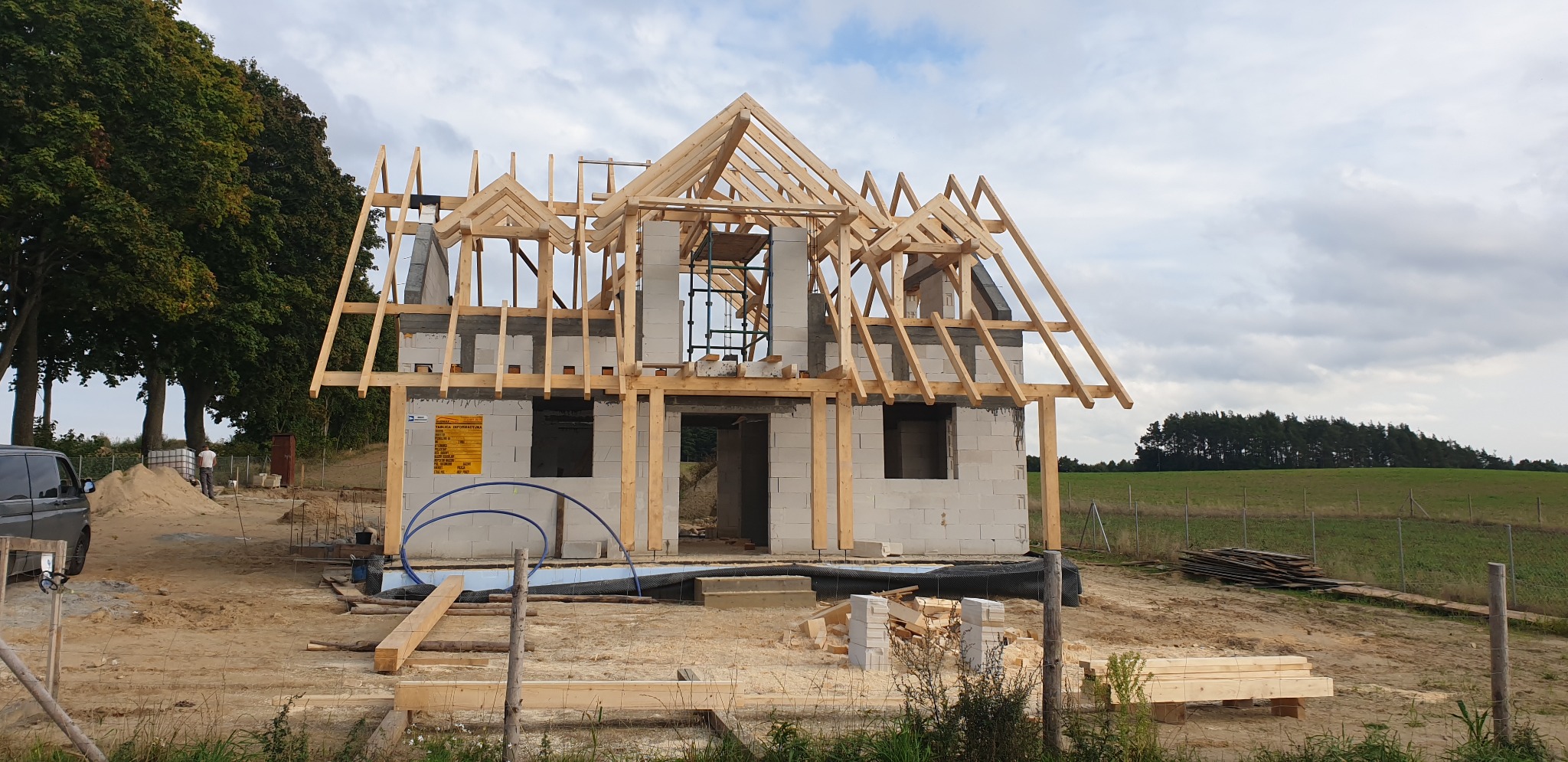 Surowy stan domu szkieletowego z widoczną konstrukcją dachu, bloczkami betonowymi i drewnianymi belkami na tle zielonego pola i pochmurnego nieba.