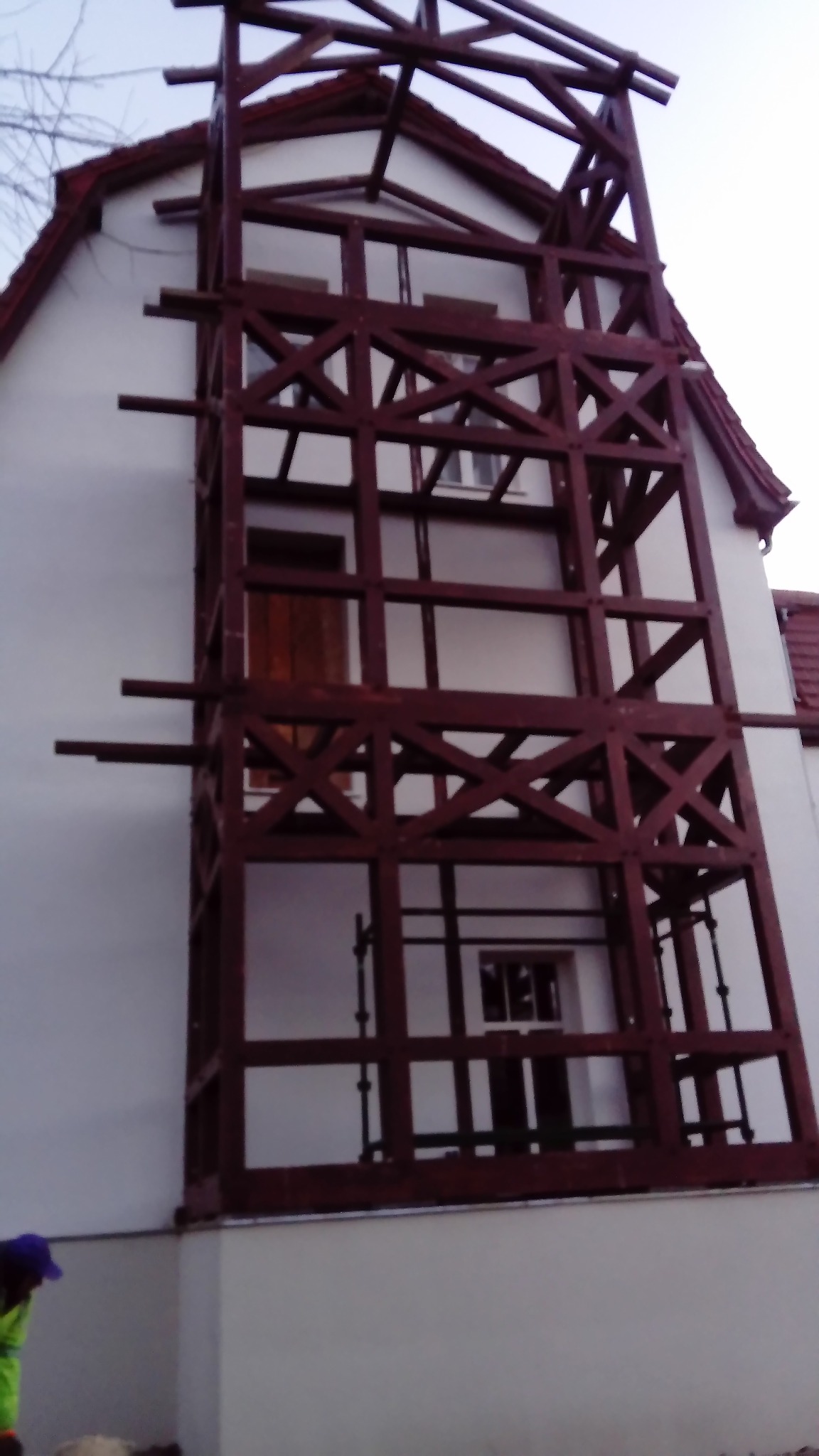 Dom w trakcie budowy z widoczną drewnianą konstrukcją szkieletową, częściowo obudowany, z widocznymi oknami i drzwiami, uchwycony z dołu.