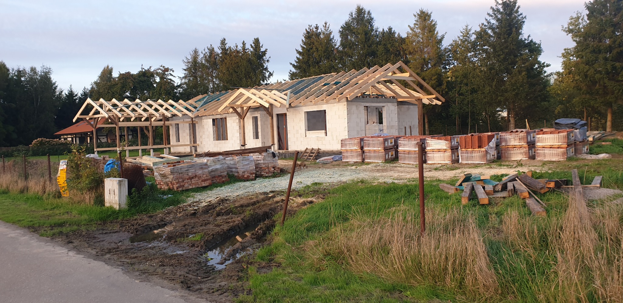 Budowa domu w stanie surowym otwartym: ściany z bloczków, drewniana konstrukcja dachu bez pokrycia, palety z dachówką obok budynku.