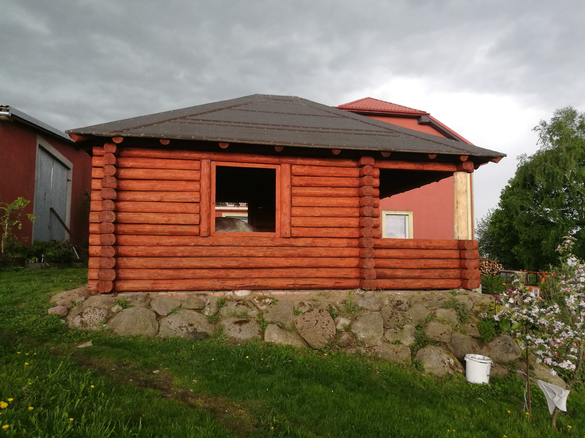 Drewniana altana z bali, posadowiona na kamiennej podmurówce, widoczna na tle pochmurnego nieba i zabudowań w odcieniach czerwieni.