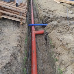 ATL Adam Grobelny - Głęboki wykop w ziemi z pomarańczową rurą kanalizacyjną i niebieskim karbowanym przewodem ułożonymi wzdłuż dna; widoczne ślady pracy i świeżo wykopana ziemia.