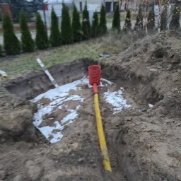Wykonany wykop pod studnię chłonną z widoczną rurą drenarską w kolorze żółtym i pomarańczową studzienką, zabezpieczony geowłókniną, na tle posesji z tujami.