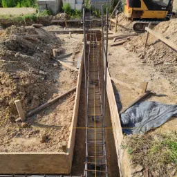 Wykop z deskowaniem i zbrojeniem pod fundament, widoczny mini-koparka, w tle budynek mieszkalny i palety z bloczkami.