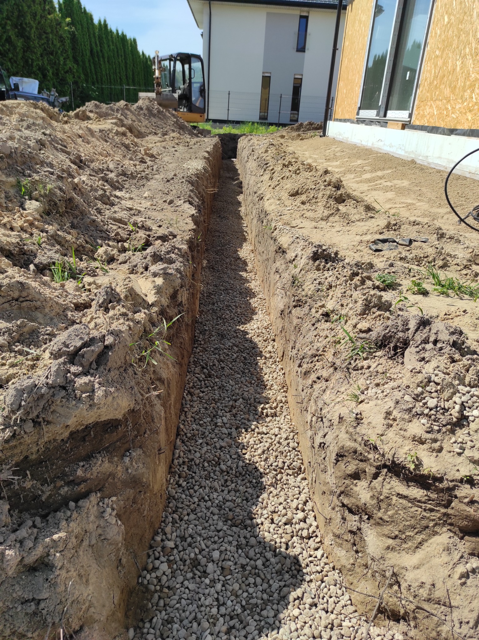Wykopany rów wypełniony drobnym żwirem, widoczny fragment budynku z oknem i koparka w tle.