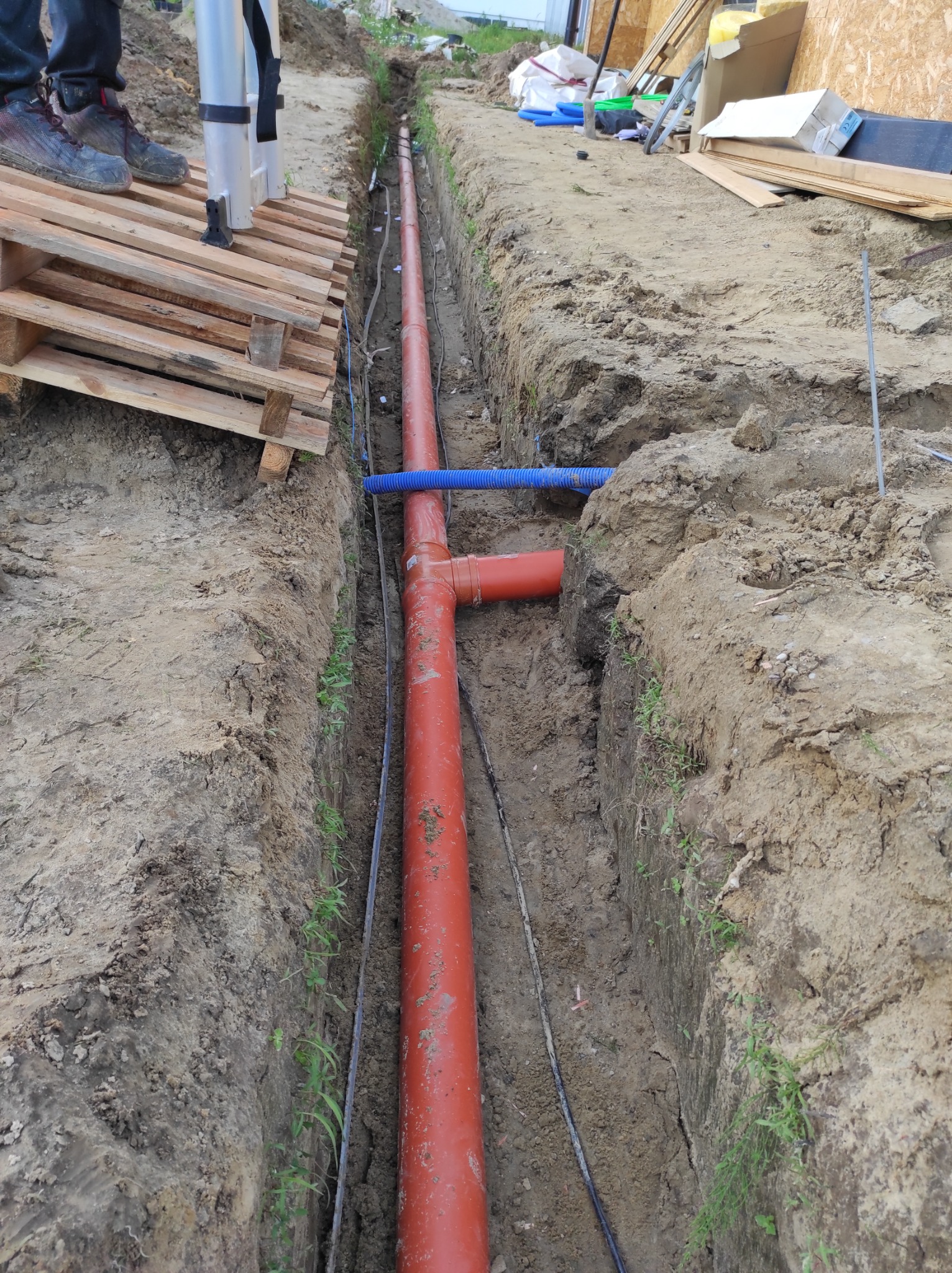 Głęboki wykop w ziemi z pomarańczową rurą kanalizacyjną i niebieskim karbowanym przewodem ułożonymi wzdłuż dna; widoczne ślady pracy i świeżo wykopana ziemia.
