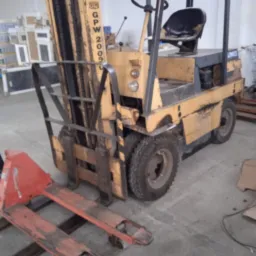 Używany wózek widłowy GPW 2009 z podczepionym czerwonym wózkiem paletowym na betonowej posadzce magazynu.