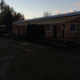 MalBud - Budynek z drewnianą konstrukcją zadaszenia w trakcie budowy, widoczne elementy mocujące i surowe drewno, w tle zachodzące słońce.