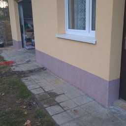 MalBud - Świeżo pomalowana elewacja budynku z widocznym oknem i otwartym garażem, dolna część ściany w kolorze fioletowym, górna w beżowym odcieniu, narzędzie malarskie leży obok.