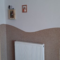 MalBud - Pokój w Konarzynach z dekoracyjną, falistą strukturą na ścianie i białym grzejnikiem panelowym; widoczna także szafka z kluczami i obrazek na ścianie.