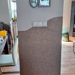 MalBud - Fragment ściany z nietypowym wykończeniem korkowym, widoczne włączniki światła, ramki ze zdjęciami na górze ściany, fragment krzesła i podłogi.