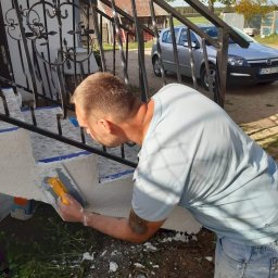 MalBud - Mężczyzna z tatuażem na ramieniu nakłada żółtą pacą gładź na betonowe schody zewnętrzne z metalową balustradą, widoczny samochód i budynek w tle.