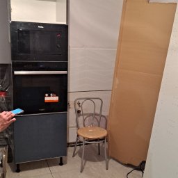 MalBud - Montaż zabudowy kuchennej: wbudowany piekarnik i mikrofalówka Amica w szafce, obok krzesło na tle płytek ściennych i kartonów, widoczny fragment dłoni z telefonem, podłoga wyłożona płytkami.