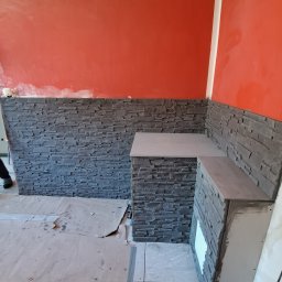 MalBud - Ściana wyłożona szarą kamienną okładziną, tworząca półkę w rogu pokoju z pomarańczową ścianą w trakcie malowania, podłoga zabezpieczona kartonem.