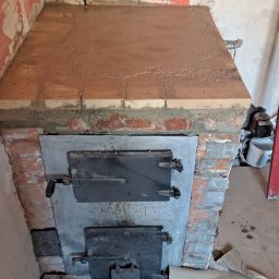 MalBud - Fragment nowo wybudowanego pieca kaflowego z metalowymi drzwiczkami, widoczny w trakcie prac wykończeniowych pomieszczenia, z surowymi ścianami i podłogą zabezpieczoną kartonem.