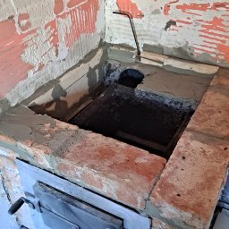 MalBud - Murowany piec w trakcie budowy, z widocznymi cegłami, zaprawą i metalowymi drzwiczkami, na tle częściowo otynkowanej ściany z resztkami farby.