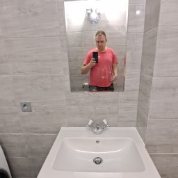 MalBud - Łazienka po remoncie z umywalką na niebieskiej szafce, lustro odbija mężczyznę robiącego zdjęcie telefonem, ściany wyłożone płytkami imitującymi drewno.