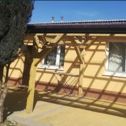 MalBud - Żółta elewacja domu z drewnianą pergolą rzucającą cienie, widoczne okna z białymi ramami i mały składany stolik.