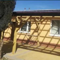 Ż&oacute;łta elewacja domu z drewnianą pergolą rzucającą cienie, widoczne okna z białymi ramami i mały składany stolik.