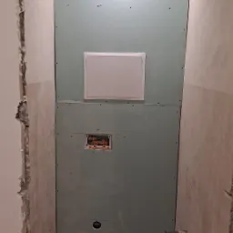 Wnętrze łazienki w trakcie prac remontowych: zamontowane płyty gipsowo-kartonowe na ścianach, widoczne przyłącza wodno-kanalizacyjne, otwór rewizyjny i surowe wykończenie ścian.