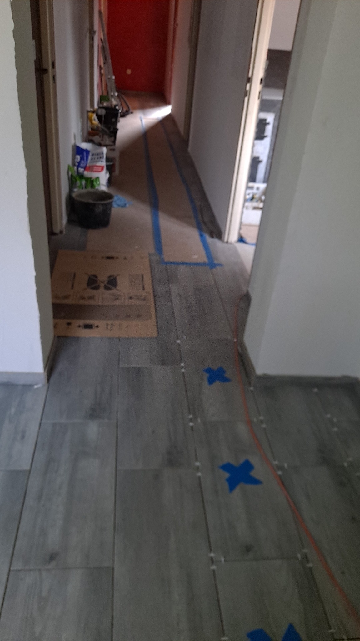 Korytarz w trakcie układania szarych płytek imitujących drewno, widoczne krzyżyki dystansowe, niebieska taśma wyznaczająca obszar roboczy, wiadro z zaprawą i narzędzia w tle.