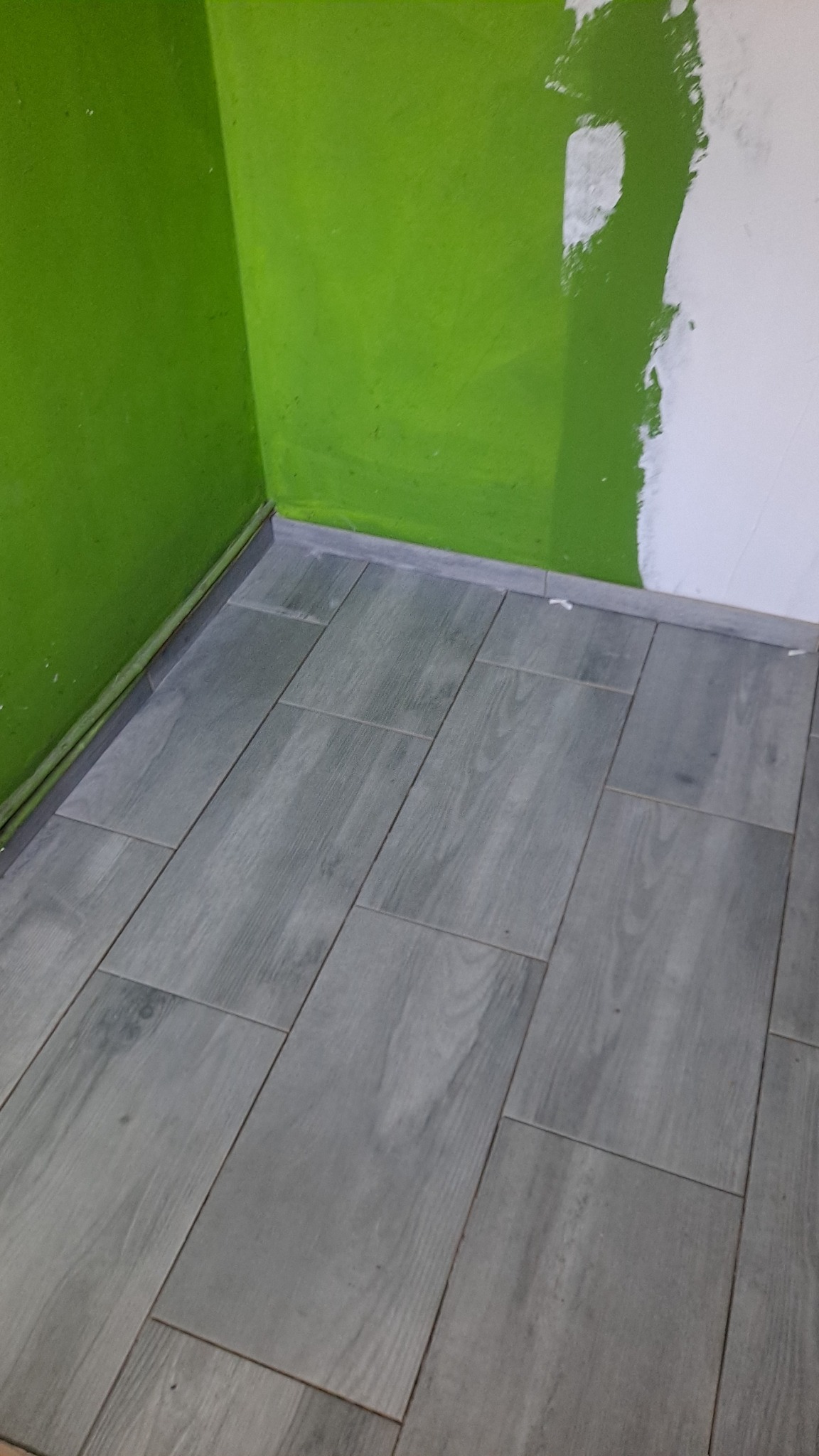Ułożona szara podłoga z płytek imitujących drewno, widoczne zielone i białe ściany w narożniku pomieszczenia, listwa przypodłogowa w kolorze płytek.