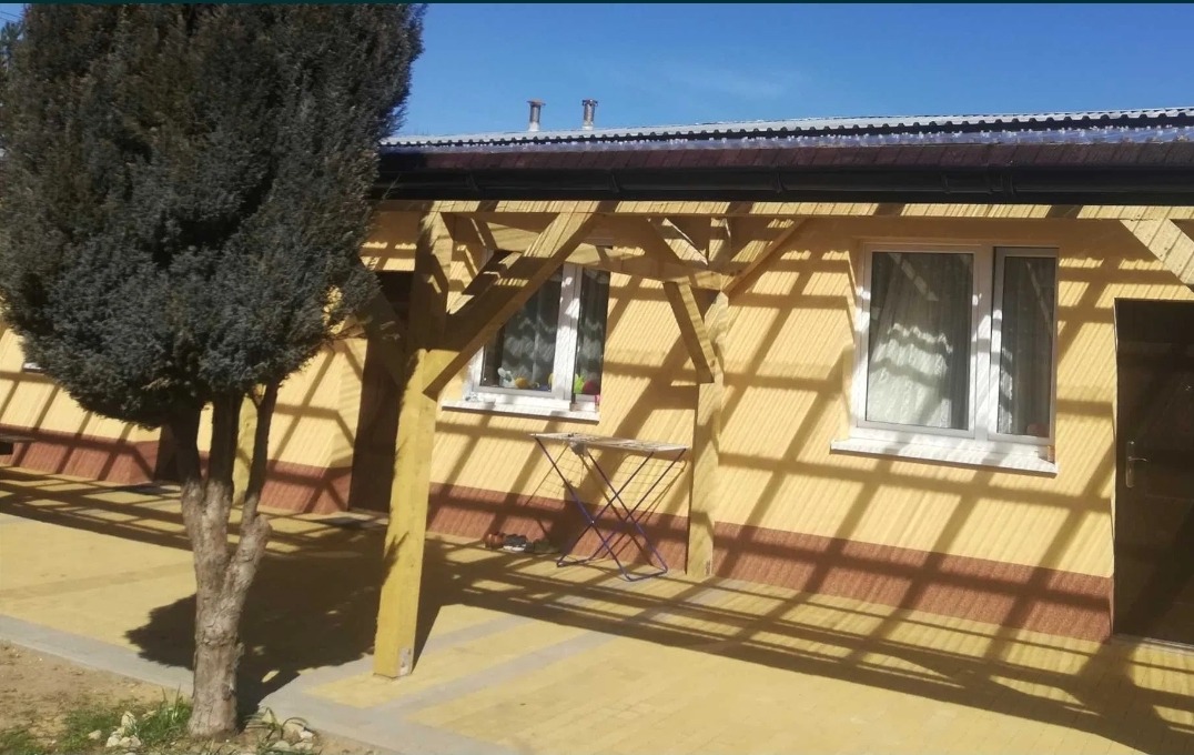 Żółta elewacja domu z drewnianą pergolą rzucającą cienie, widoczne okna z białymi ramami i mały składany stolik.