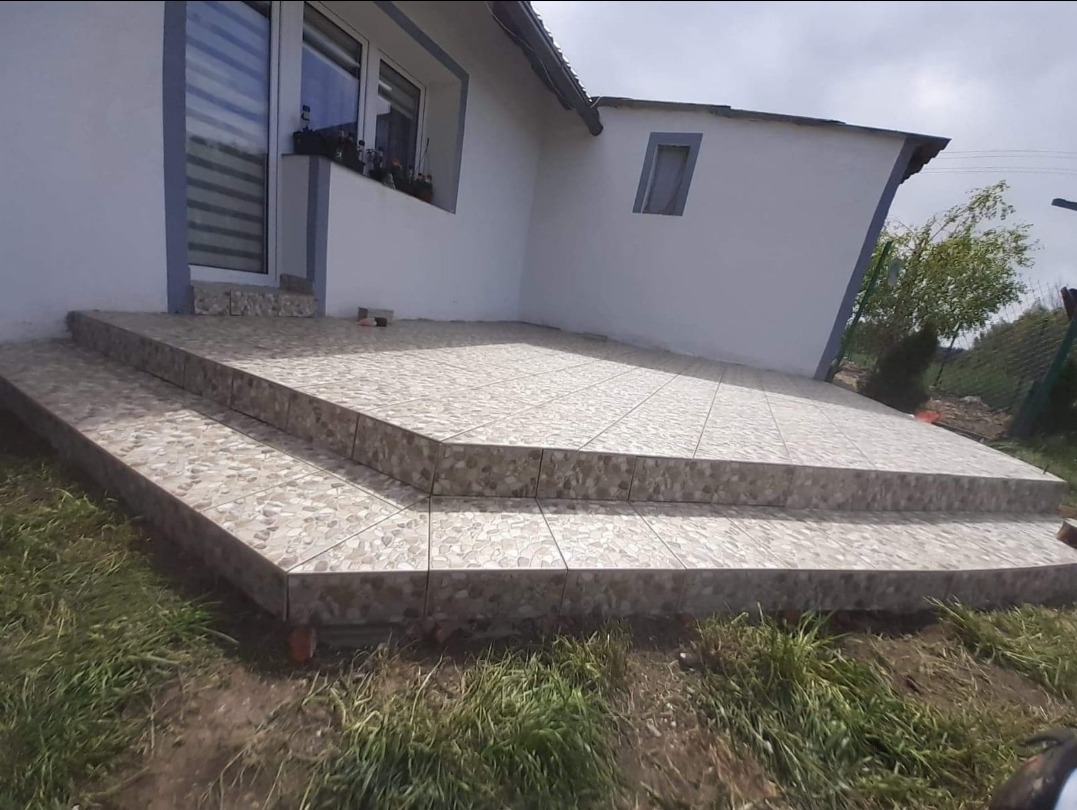 Wykończone płytkami schody wejściowe do domu, z widoczną elewacją budynku i fragmentem ogrodu.