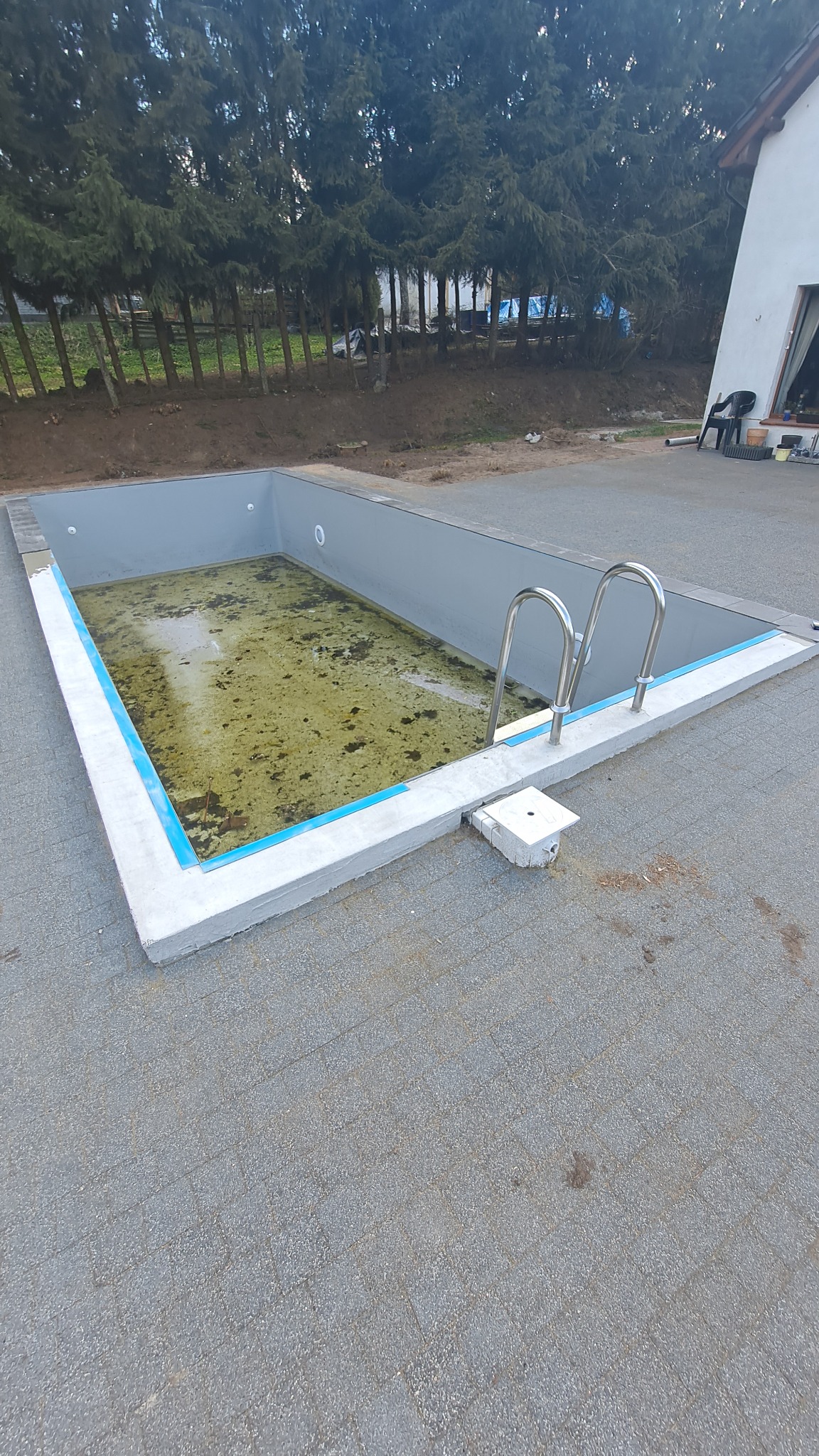 Napełniony brudną wodą basen w trakcie czyszczenia, z metalową drabinką, otoczony szarą kostką brukową i zielonymi drzewami w tle.