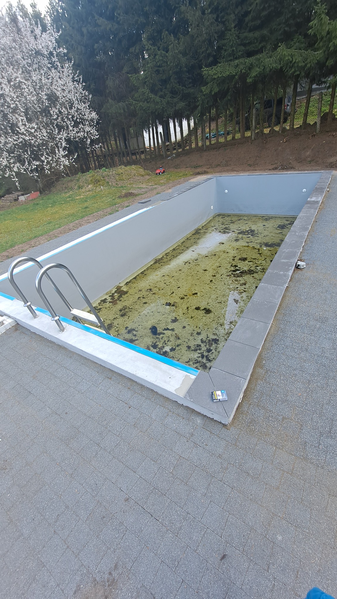 Napełniony brudną wodą basen w trakcie czyszczenia, z metalową drabinką i wyłożony szarymi płytami, widoczny pakiet papierosów na obrzeżu.