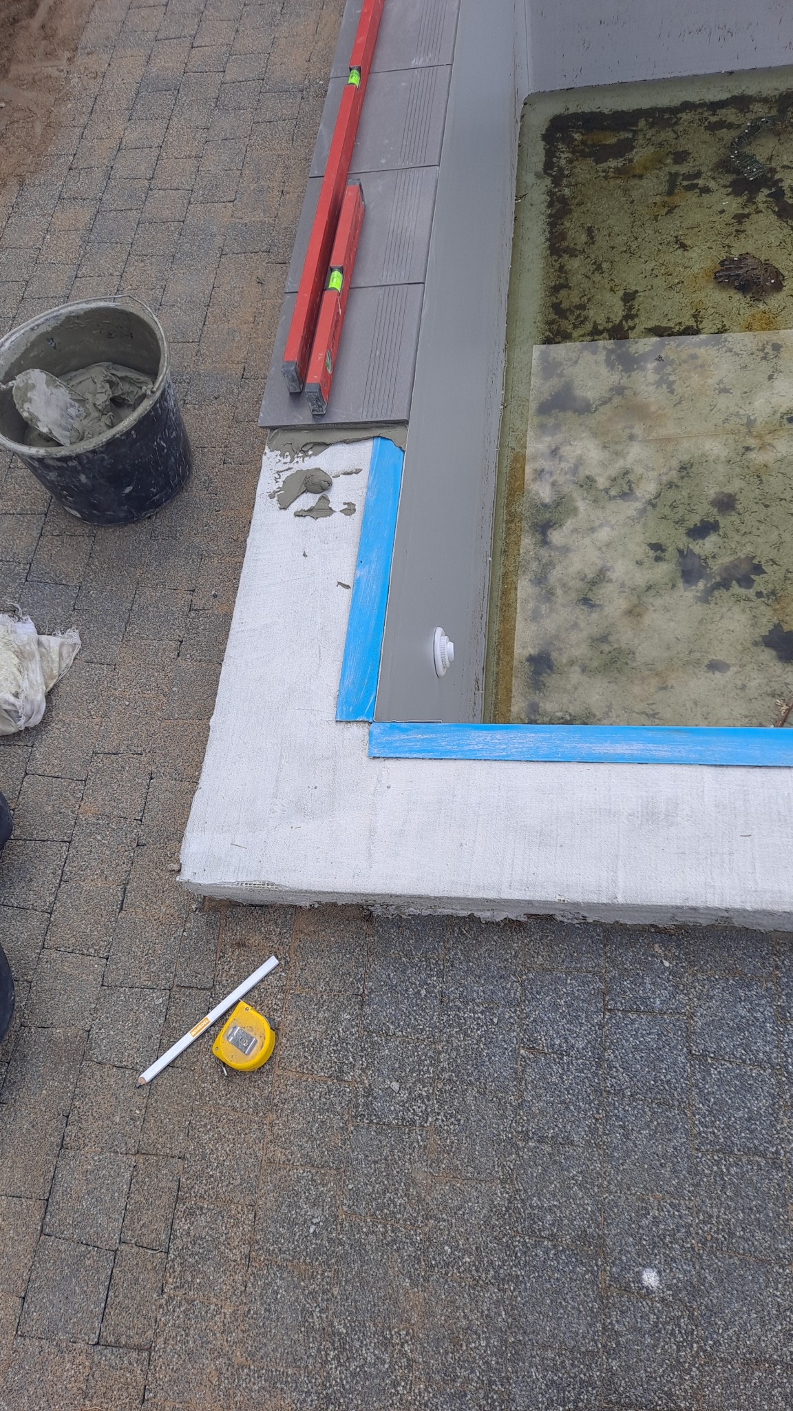 Budowa lub remont basenu: widoczny fragment krawędzi basenu z nałożonymi płytkami, poziomice, wiadro z zaprawą, miarka i ołówek na kostce brukowej.