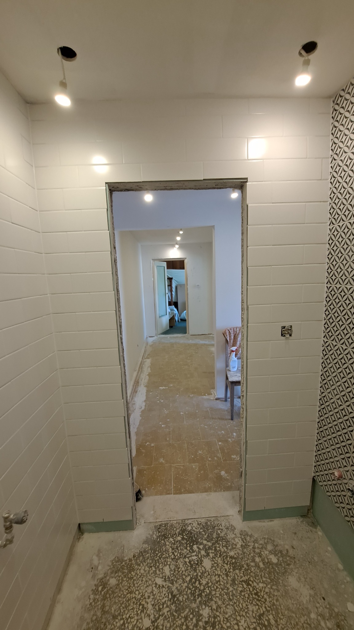 Łazienka w trakcie remontu: białe, prostokątne płytki na ścianach, wzorzysta ściana, widoczne przygotowania do położenia podłogi, surowe wykończenie framugi drzwiowej, widok na dalszą część...