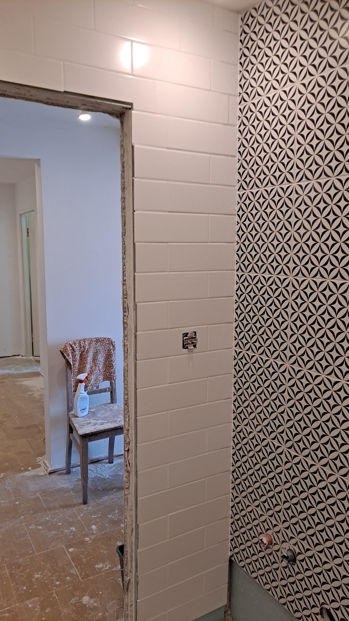 Świeżo ułożone, białe, prostokątne płytki na ścianie łazienki, kontrastujące z czarno-białą ścianą w geometryczne wzory. Widoczna puszka elektryczna bez osprzętu, a w tle krzesło z butelką środka...