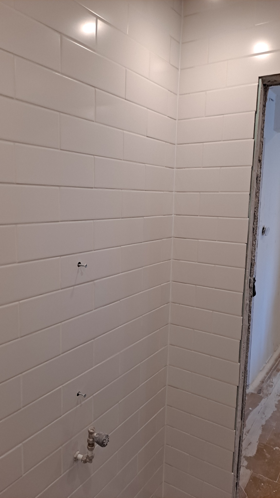 Świeżo położone, błyszczące, białe, prostokątne płytki na ścianie w rogu pomieszczenia, widoczne wystające śruby montażowe i zawór, surowy otwór drzwiowy w tle.