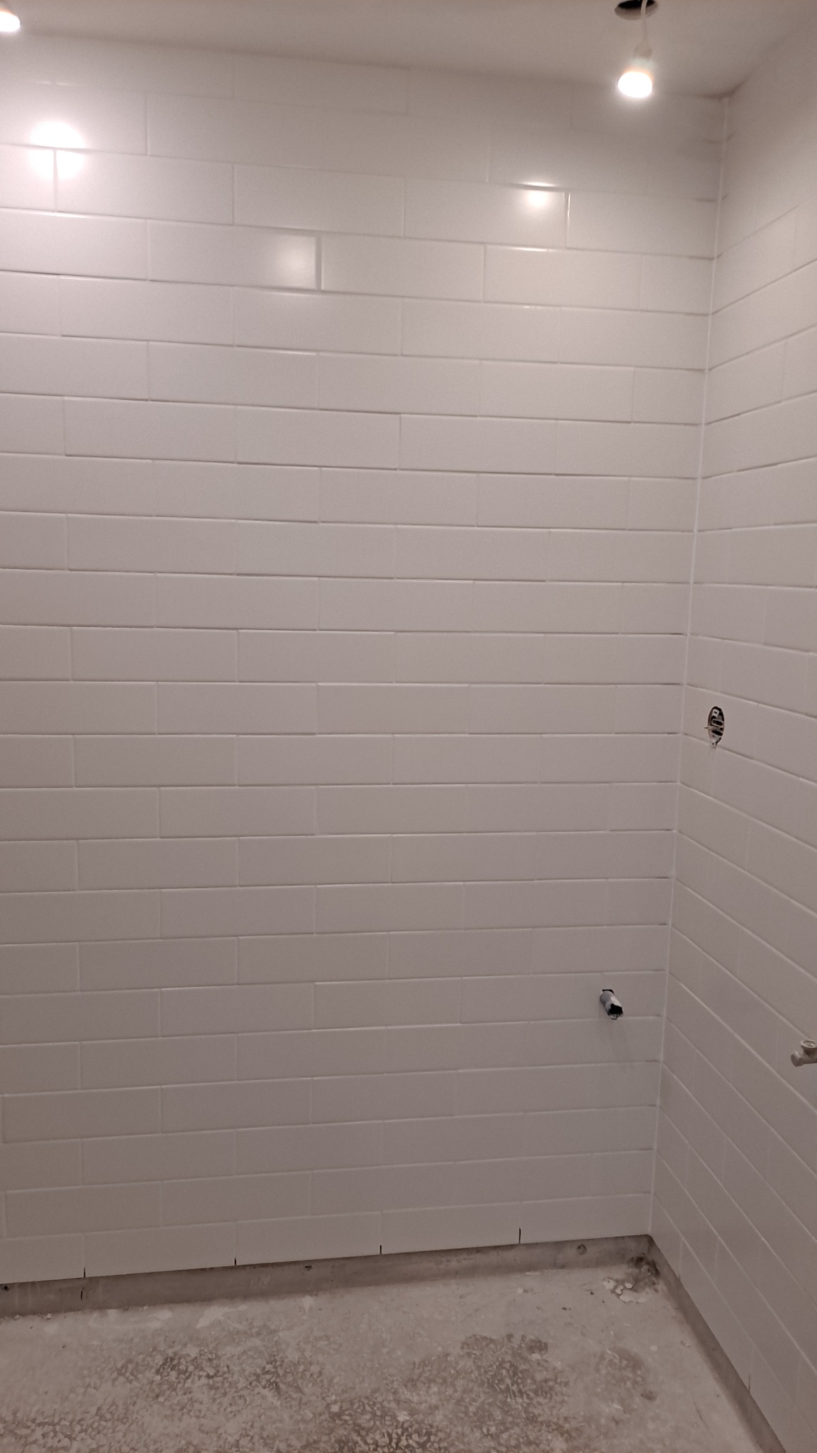 Ściana w trakcie układania białych, prostokątnych płytek, widoczne punkty mocowania armatury, surowa podłoga przed fugowaniem.