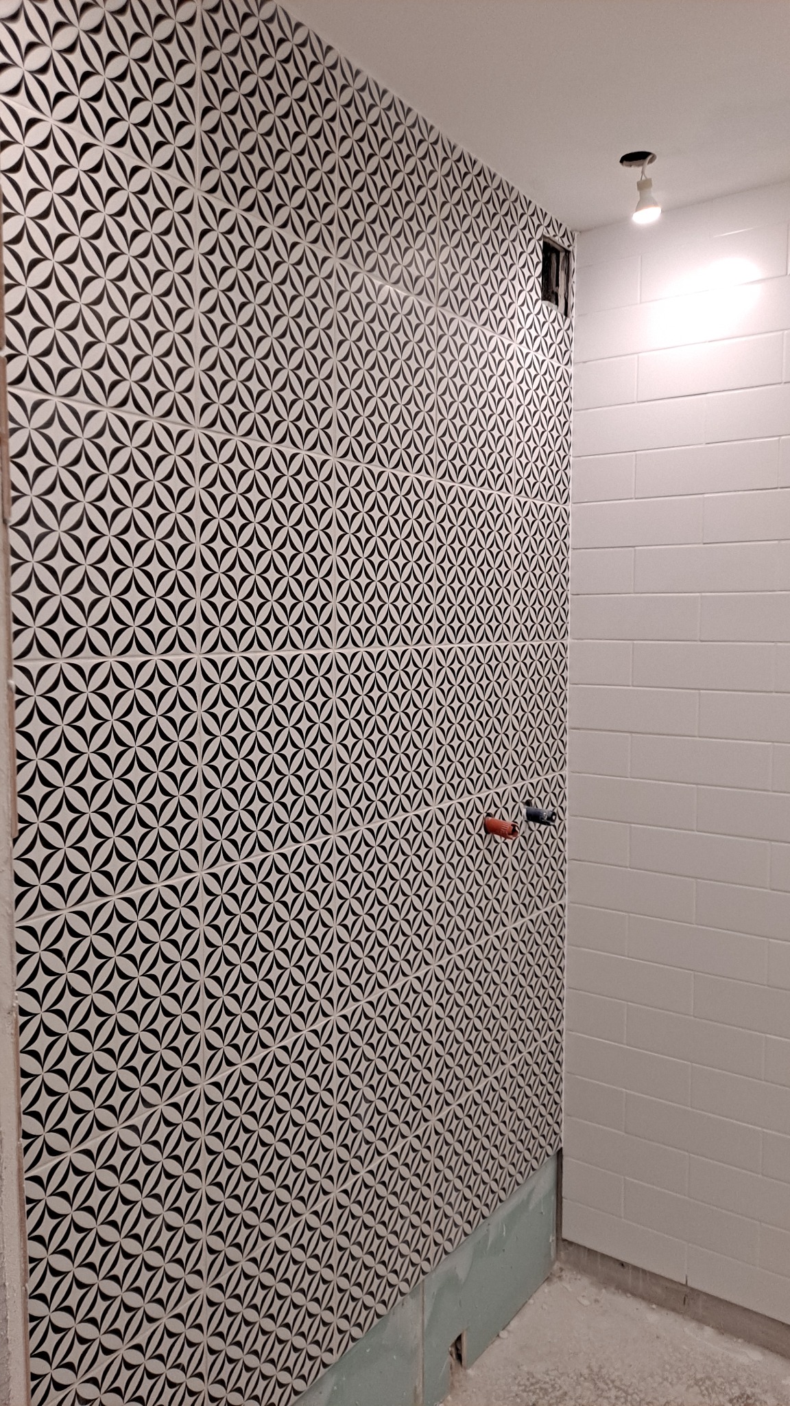 Ściana łazienki wyłożona białymi płytkami z czarnym wzorem geometrycznym, sąsiednia ściana wyłożona białymi prostokątnymi płytkami, widoczne niedokończone wykończenie podłogi i wystające rury.