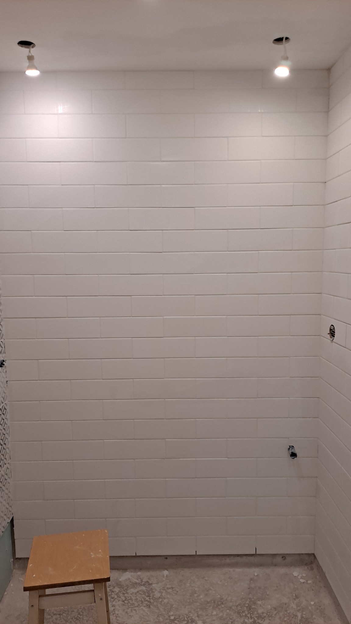 Ściana łazienki wyłożona białymi, prostokątnymi płytkami, z widocznymi punktami oświetleniowymi na suficie i drewnianym stołkiem na podłodze w trakcie prac wykończeniowych.