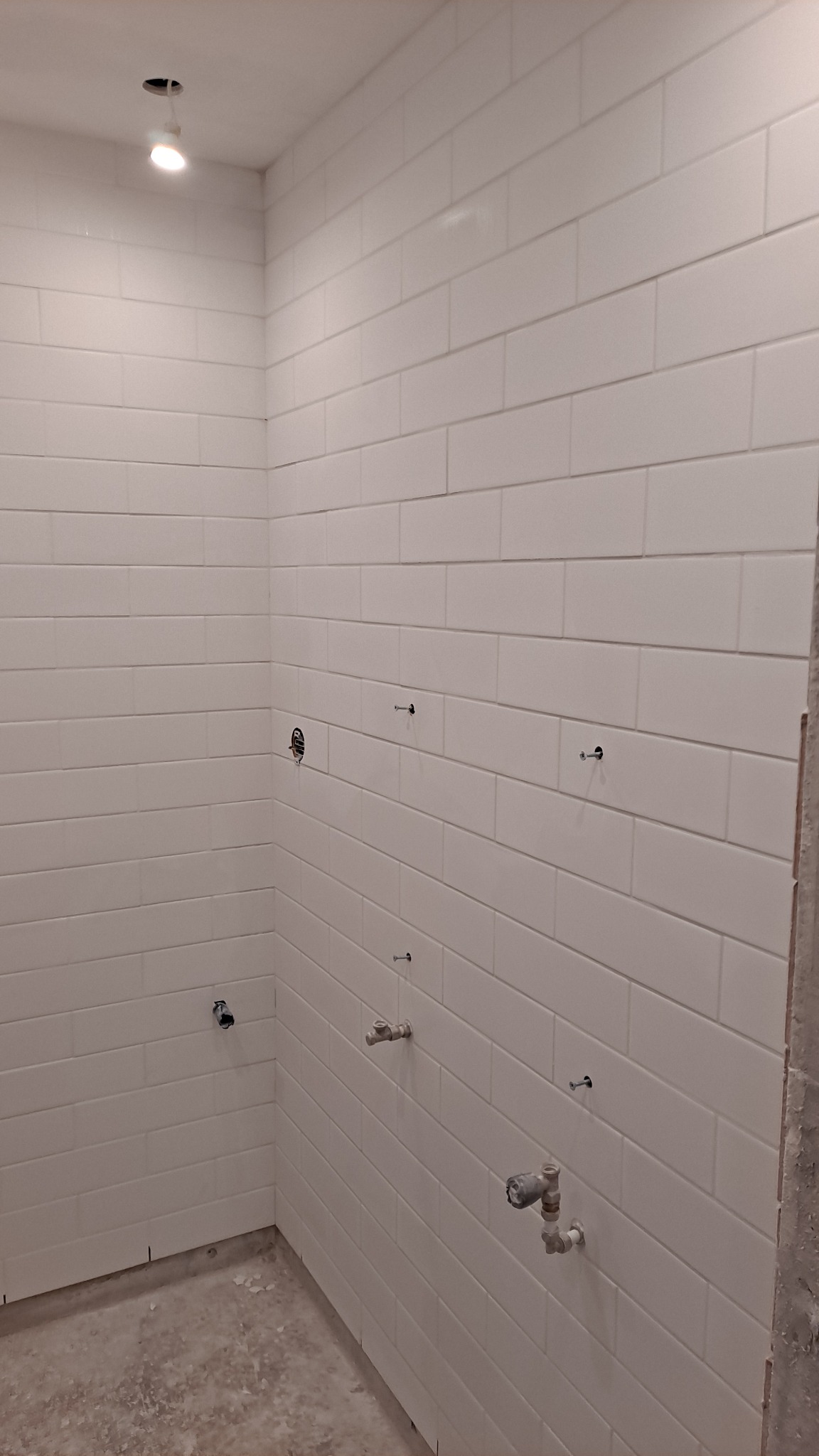 Ściana łazienki wyłożona białymi, prostokątnymi płytkami w trakcie montażu armatury, widoczne wystające przyłącza wodne i otwór na oświetlenie w suficie.