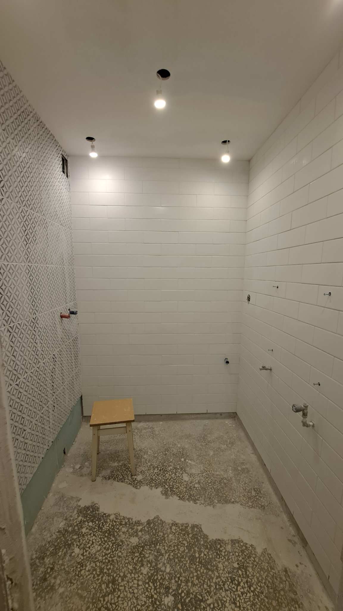 Wnętrze łazienki w trakcie remontu, z częściowo położonymi białymi płytkami na ścianach i wzorzystymi płytkami na jednej ścianie, widoczne wystające rury instalacji wodnej i elektrycznej,...